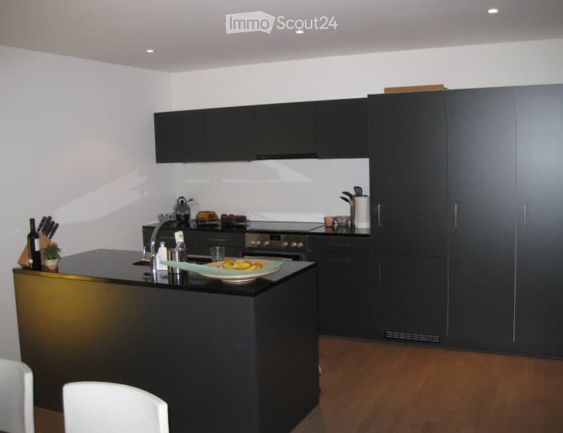 Modern kitchen with black cabinets, a central island, and stainless steel appliances. The island features a sink, faucet, and decorative items including a bowl of fruit and a bottle. The floor is wooden, and the walls are white.