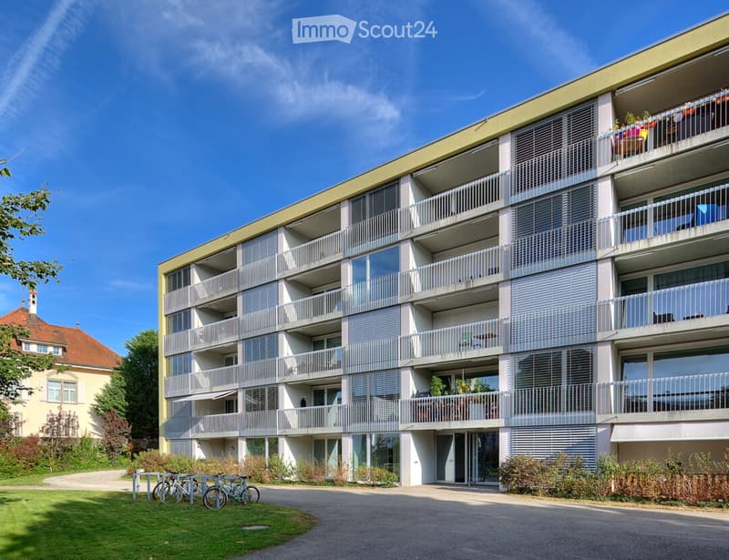 Modern multi-story apartment building with balconies, large windows, and a green lawn area with bicycles.