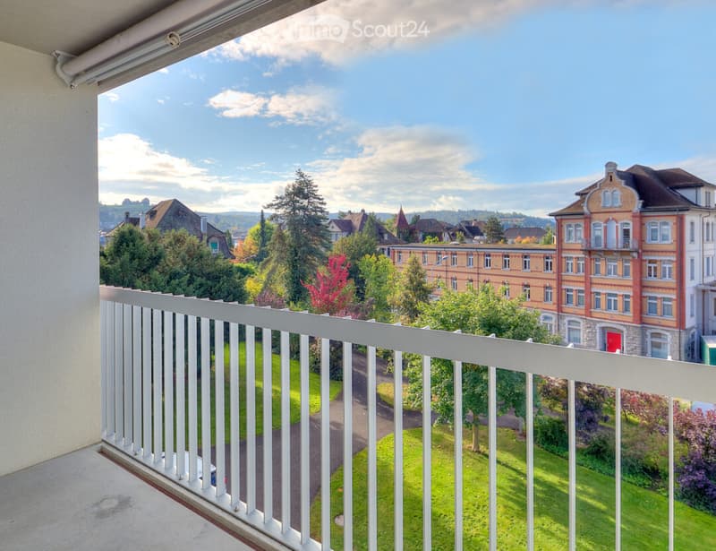 Balcony with white railing overlooking a green lawn, trees, and a multi-story building in the background.