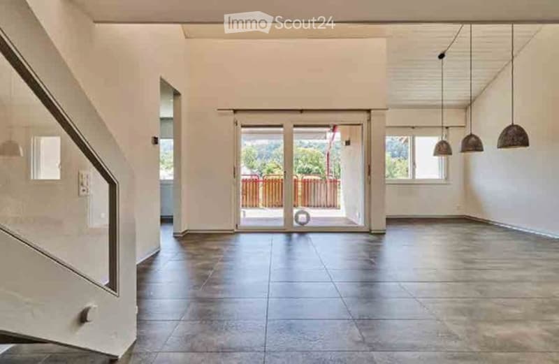 Empty, modern hallway with a staircase on the left, large glass doors leading to an outdoor area, and pendant lights hanging from the ceiling.