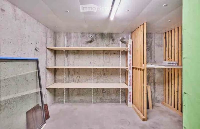 Empty room with exposed concrete walls and floors, built-in wooden shelving unit, and unfinished wooden framing on one side.