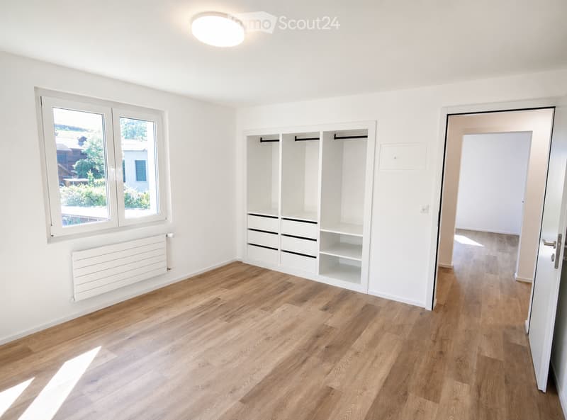 Empty room with light wooden flooring, white walls, built-in closet with shelves and drawers, large windows with white frames, and a white radiator under the windows.