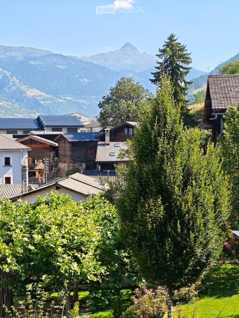 A picturesque village scene with traditional houses, lush greenery, and a backdrop of mountain peaks under a clear blue sky.