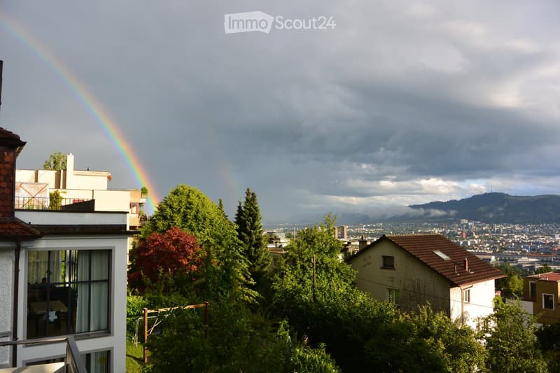 A multi-story residential building with a balcony overlooking a cityscape, surrounded by trees and a visible rainbow in the sky.