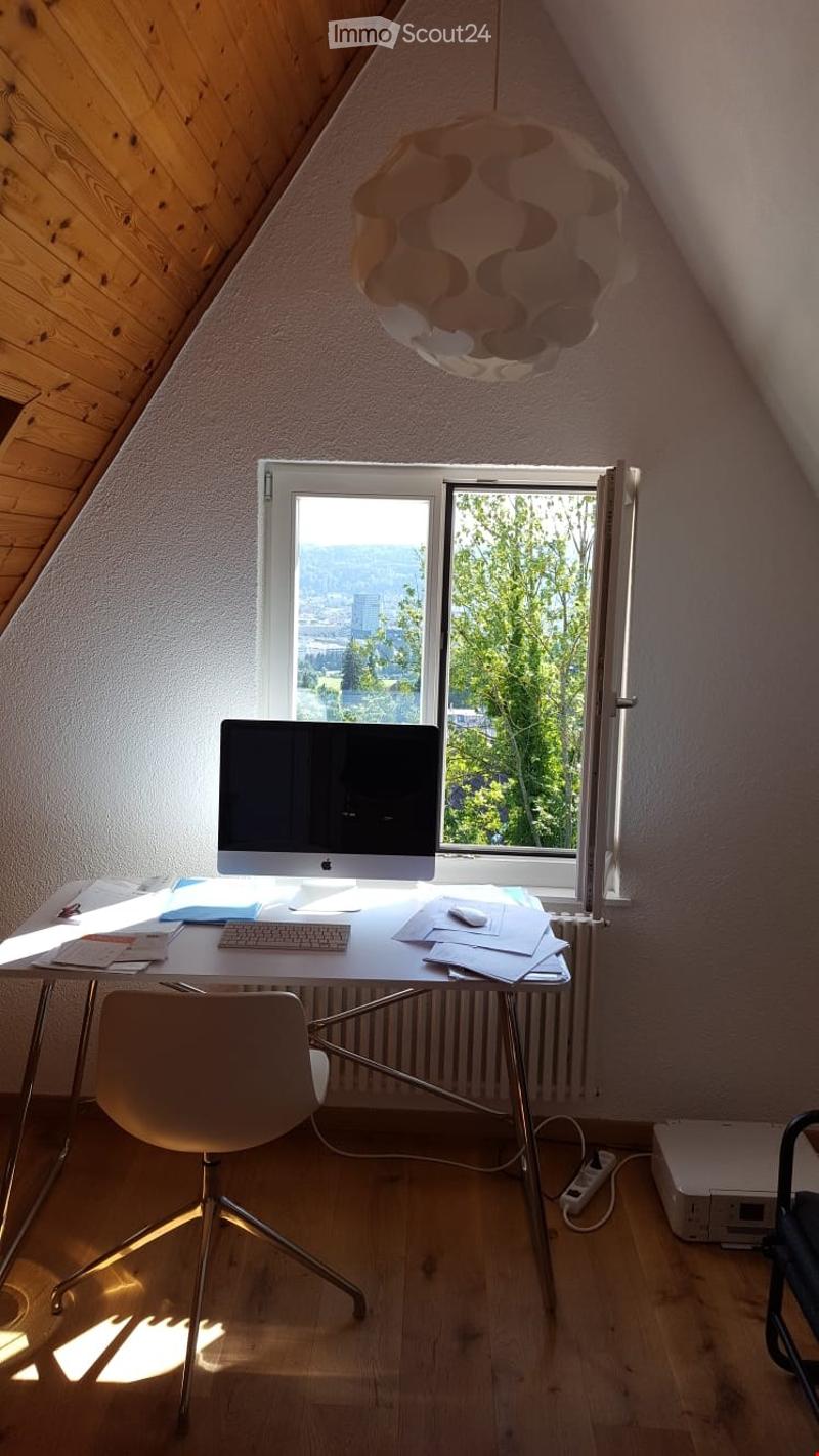 Bright attic room with wooden floors, large window offering a scenic view, modern desk setup with computer, keyboard, mouse, and chair.
