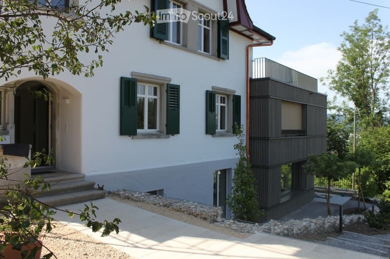 Two-story house with white exterior, green shutters, and a modern balcony on the upper level. The entrance features a small staircase leading to a dark door, and the ground floor includes a raised section with a stone border.