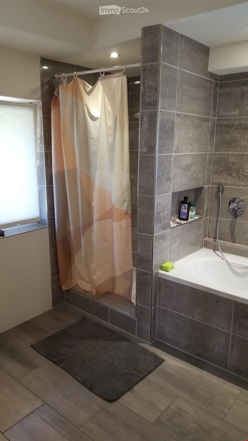 Modern bathroom with a bathtub, shower curtain, and a shelf holding toiletries. The walls are tiled in grey, and the floor is tiled in beige.