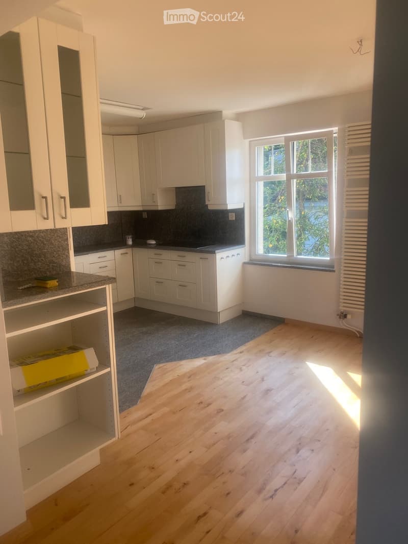 Empty kitchen with white cabinets, granite countertops, and wooden flooring. The space is clean and well-lit with natural light from a large window.