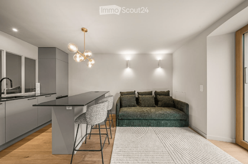 Modern, open-concept living and dining area with a grey sofa, kitchen island, and sleek bar stools. The space features contemporary lighting and a minimalist design.