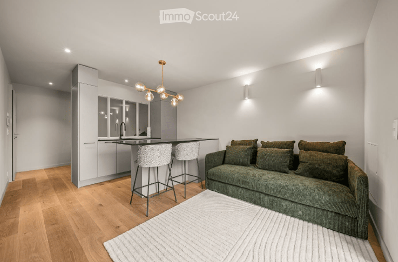 A modern, open-concept living and dining area with a green sofa, two stool chairs, a kitchen island, and sleek kitchen cabinets.
