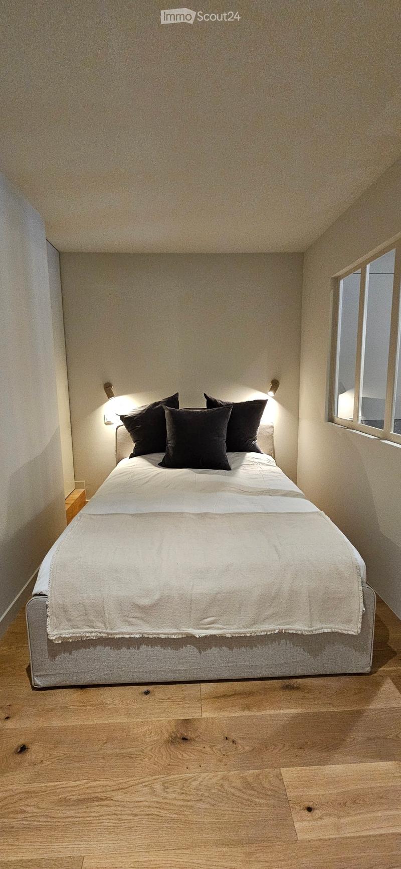 A modern bedroom with a neatly made bed, white bedding, black pillows, and a wooden nightstand. The room features light-colored walls and wooden flooring.