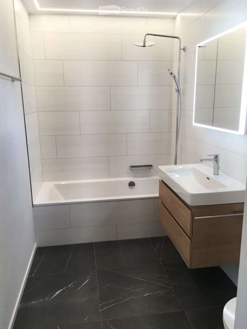 Modern bathroom with white tiles, black marble flooring, a bathtub, a sink with a wooden cabinet, and a showerhead with handheld sprayer.