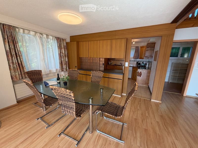 Dining area with a glass table, four wicker chairs, and a small plant on the table. The room features wooden cabinets, a window with curtains, and a view into an adjacent kitchen area with appliances.