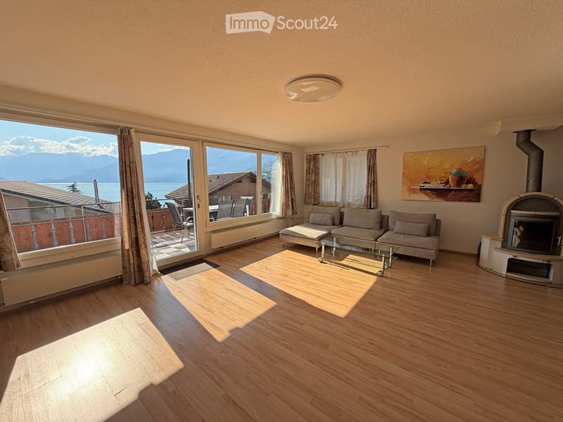 Bright living room with wooden flooring, large windows offering a scenic mountain view, a sofa, coffee table, ottoman, fireplace, and a balcony with outdoor furniture.