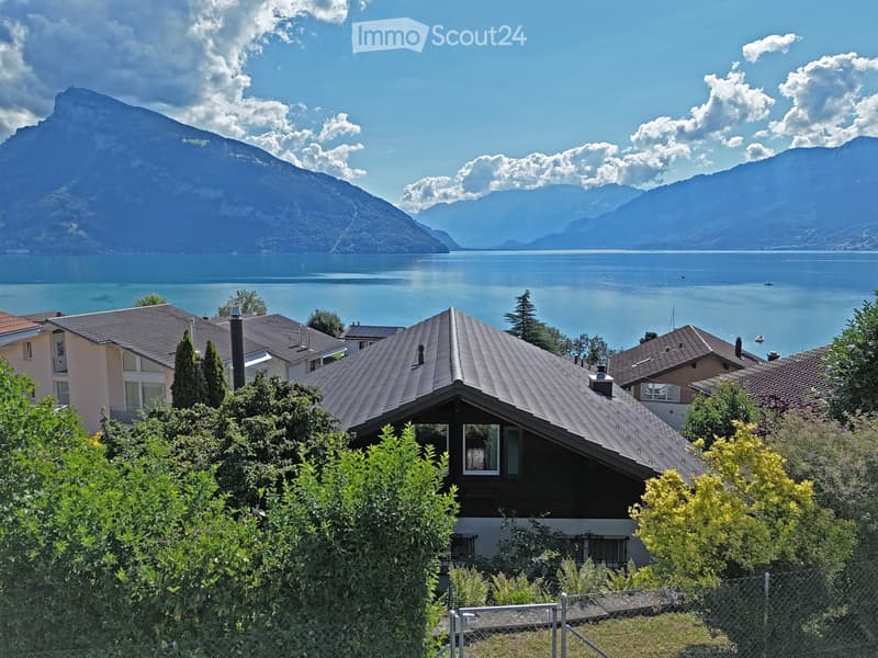 A house with a grey roof and white walls, surrounded by greenery, overlooking a lake and mountains.