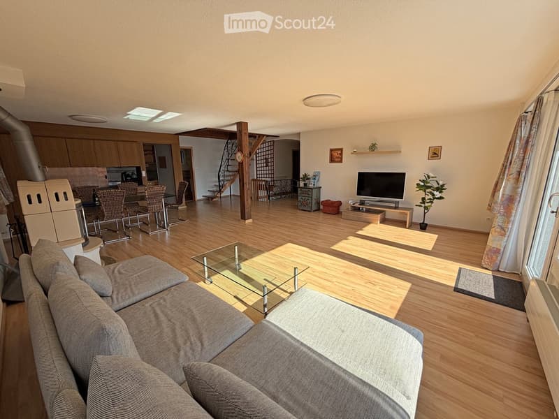 A modern living room with a grey sectional sofa, glass coffee table, flat-screen TV, dining area with chairs and table, and a refrigerator in the background. The space features wooden flooring, large windows with curtains, and decorative elements like pla