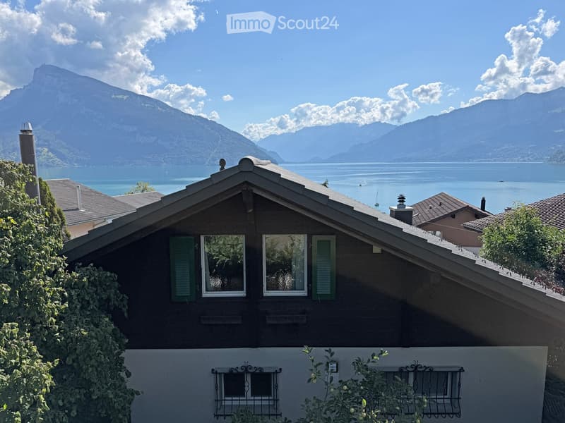 A two-story house with dark brown walls, green shutters, and a tiled roof, situated in a scenic lakeside location with mountains in the background.