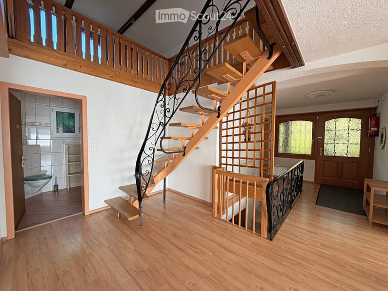 Spacious hallway with wooden flooring, a curved staircase with black wrought iron railing, and a wooden railing leading to an upper level. Features include a small table, a door, and a glimpse of a bathroom with a toilet and sink.