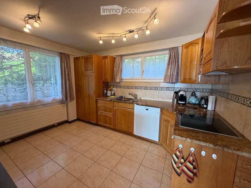 A well-lit kitchen with wooden cabinets, granite countertops, and various appliances including a dishwasher, microwave, and coffee maker. The space features tiled flooring and large windows with curtains.