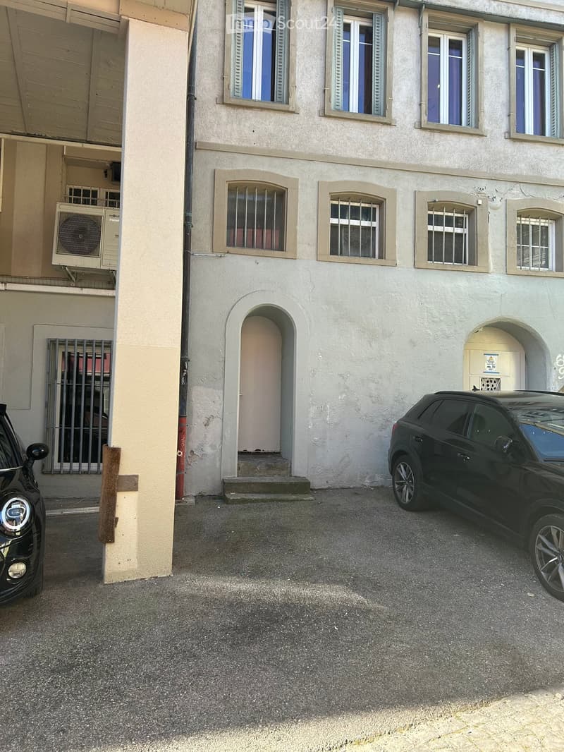A multi-story residential building with white walls, arched doorways, and multiple windows with shutters. Two cars are parked in front of the building.
