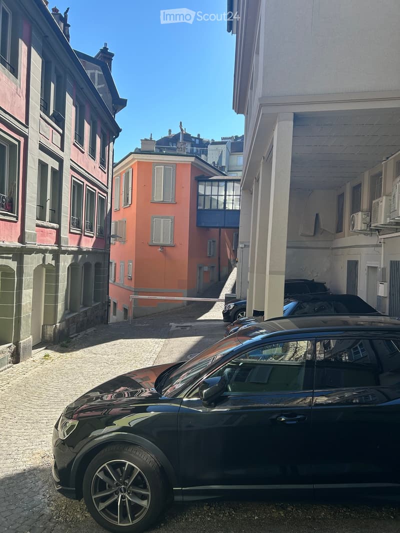 A narrow urban street lined with multi-story buildings, featuring a mix of architectural styles and parked cars.