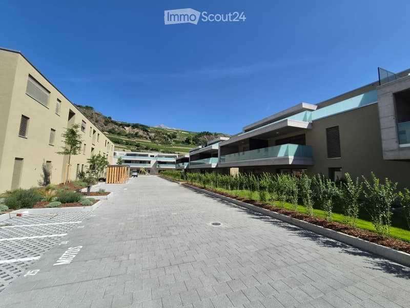 Modern residential complex with multiple buildings, balconies, landscaped gardens, and a paved walkway.