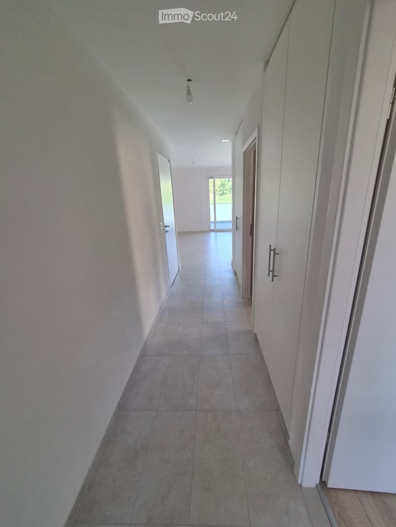 Empty hallway with tiled floor, white walls, and recessed lighting. Doors lead to other rooms, with a view of an outdoor area at the end.