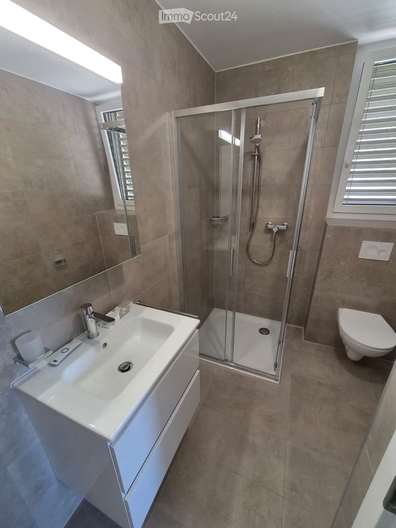 Modern bathroom with a white sink, chrome faucet, large mirror, glass-enclosed shower, and toilet. The walls and floor are tiled in a neutral tone.