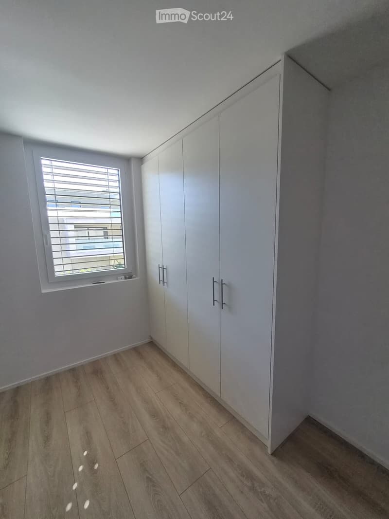 Empty room with light wooden flooring, white walls, built-in white wardrobes, and a window with blinds.