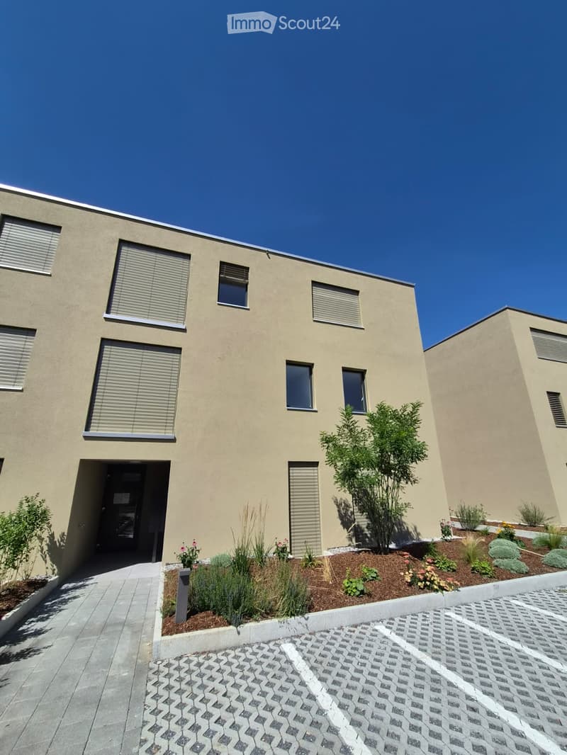 Modern two-story house with beige stucco exterior, multiple windows with blinds, small landscaped garden area with plants and flowers, and a paved driveway.