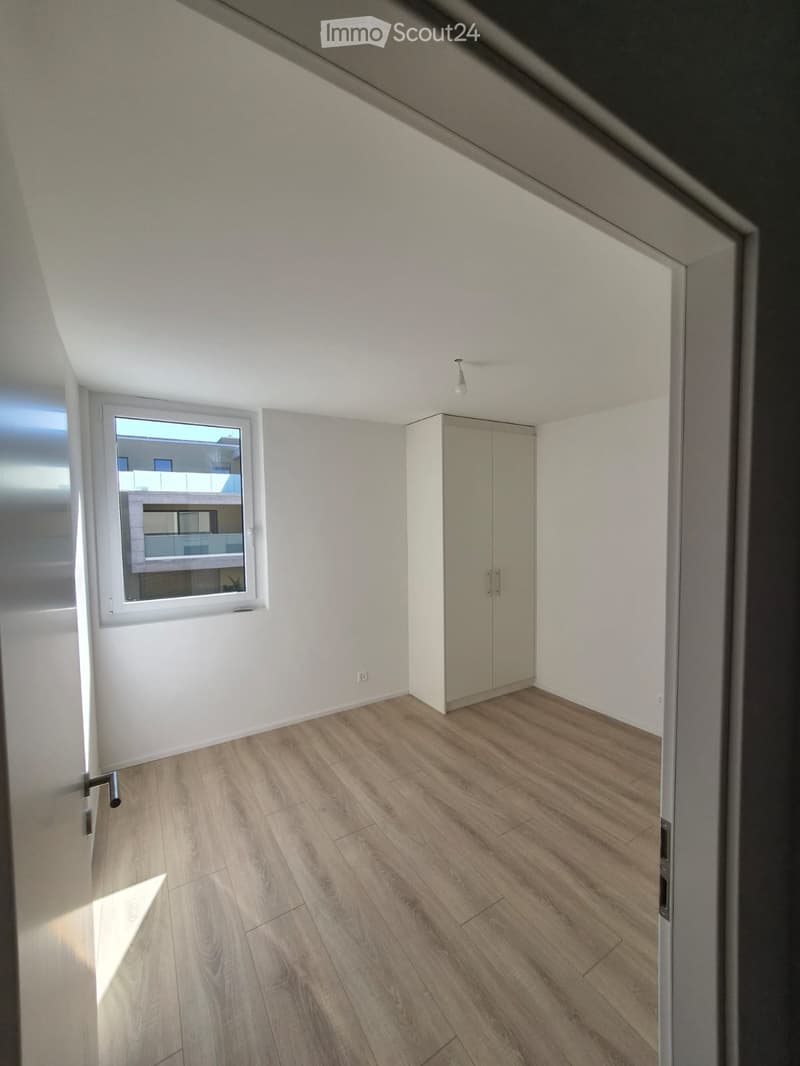 Empty room with light wooden flooring, white walls, and a small built-in closet. A window offers a view of another room with a balcony.