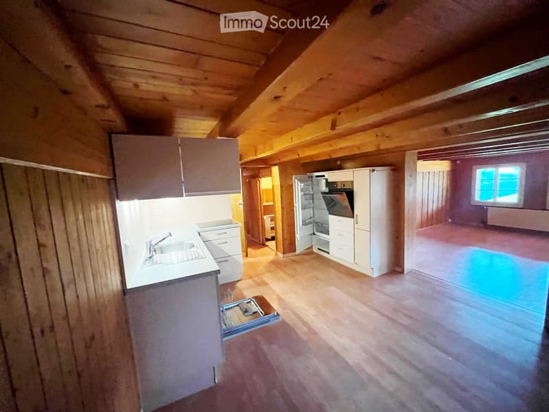 Open kitchen area with wooden beams, white cabinets, sink, faucet, refrigerator, and oven. The space is connected to a larger room with wooden flooring.