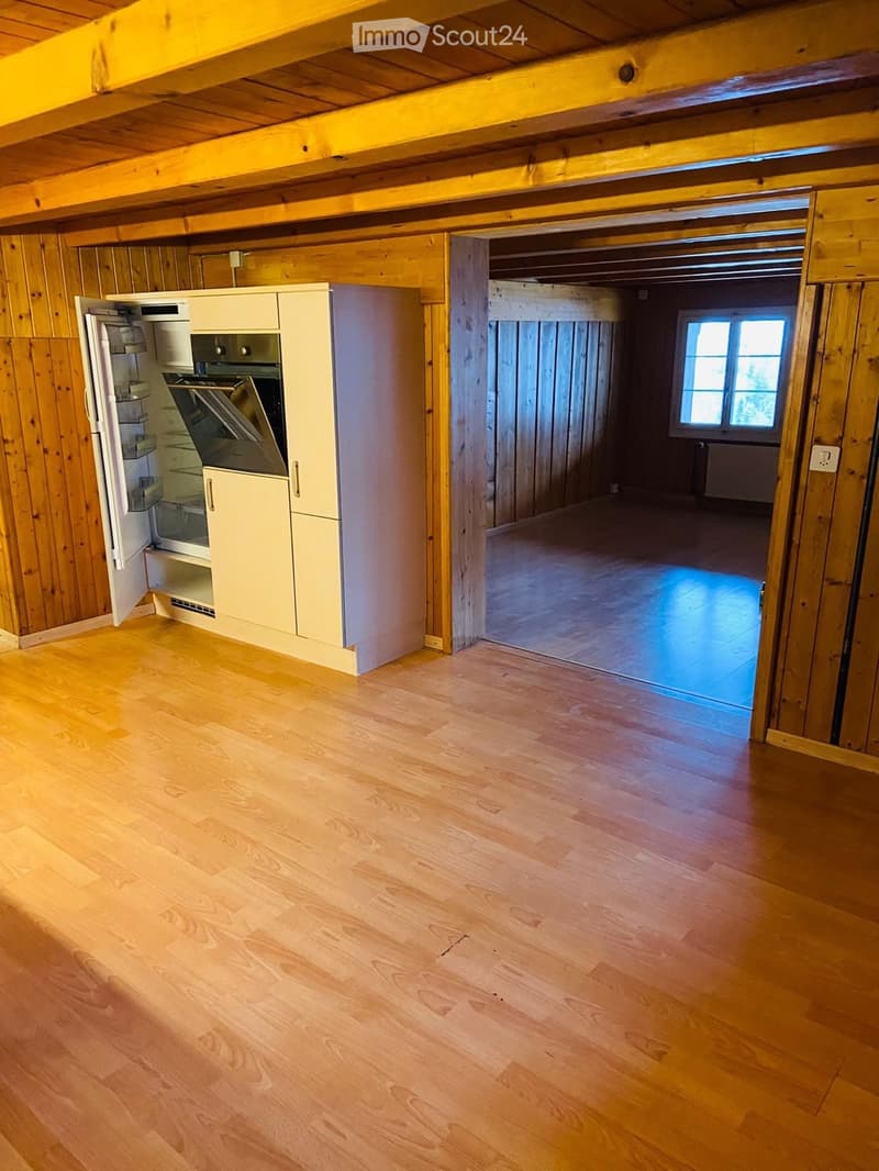 Empty room with wooden walls and floor, featuring a partially visible kitchen area with cabinets and a refrigerator on the left side. The room has exposed wooden beams on the ceiling and a doorway leading to another room with a window.