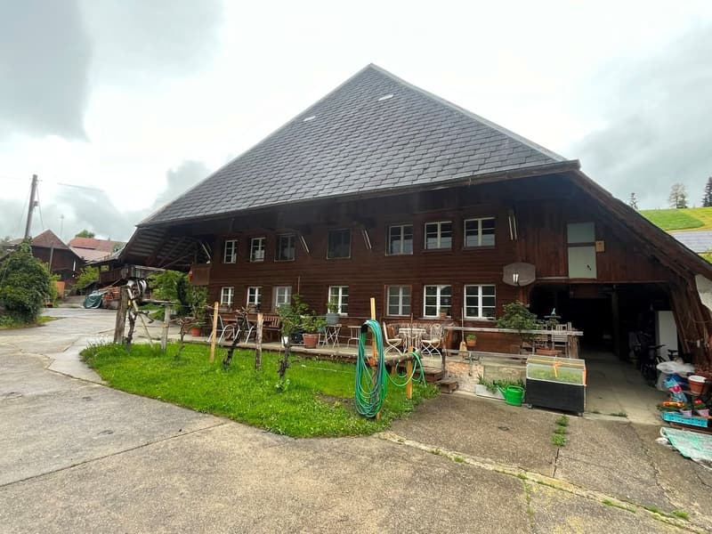 Traditional wooden house with a steep roof, multiple windows, and a spacious outdoor area with chairs, tables, and greenery.