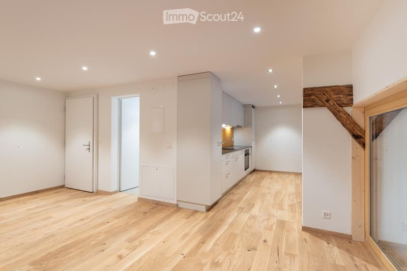 empty room with wooden floors, kitchen area, large window, wooden beam