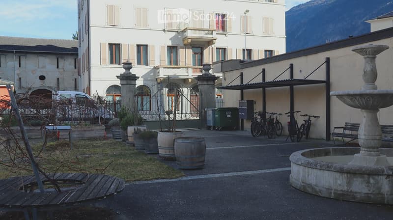 white building, multiple windows, bicycles parked, fountain, barrels, grass, mountains in the background