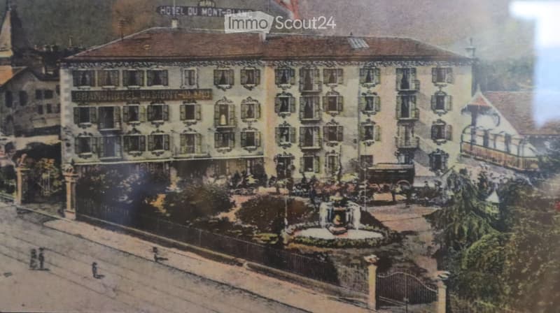 Hotel building with many windows, brown roof, garden in front, fountain, fence