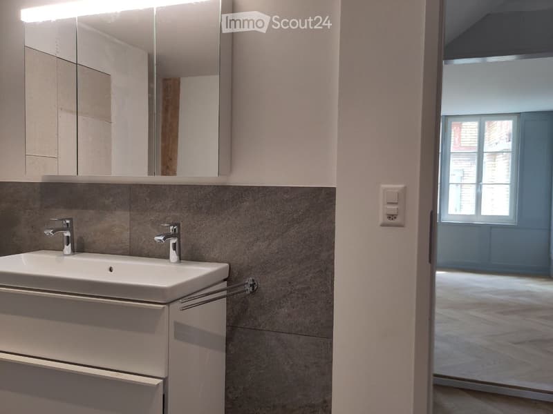 Modern bathroom with a white sink, mirrored cabinet, and grey tiled walls. The room features a sleek design with a visible doorway leading to another space.