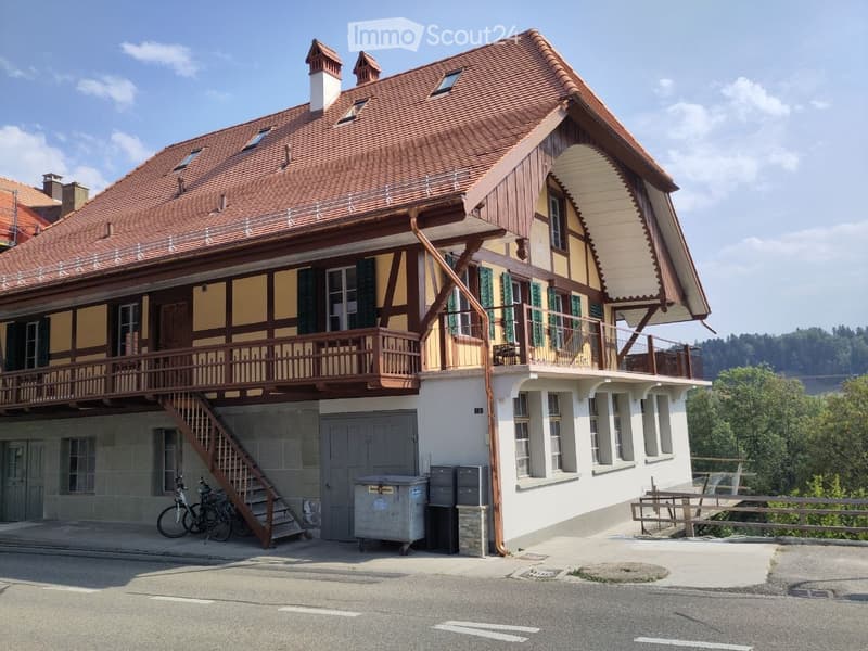 Two-story house with red-tiled roof, wooden balcony, and green shutters. Features a staircase leading to the entrance and bicycles parked nearby.