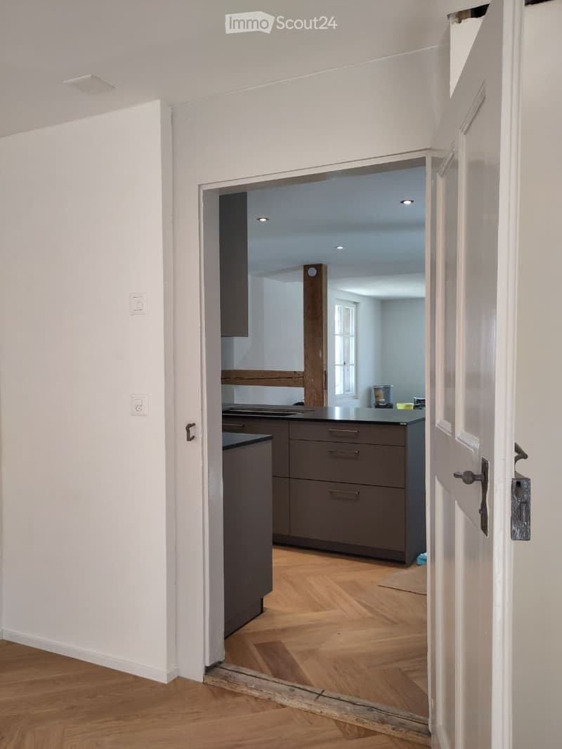 Open door leading to a kitchen with wooden floor, white walls, wooden beam, wooden cabinets, and a countertop.