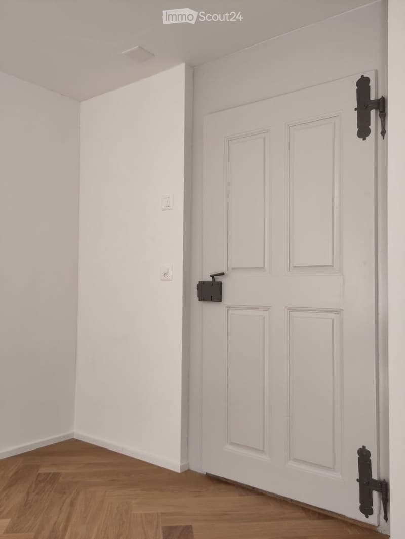 Empty room with white walls, wooden floor, white door with black handles and locks, and two white switches on the wall.