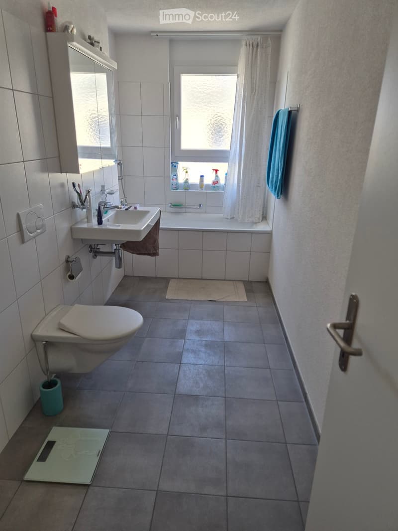 Modern bathroom with white tiles, a toilet, sink, bathtub, mirror, and cleaning supplies on the windowsill. Includes a towel, bathroom scale, and a toothbrush holder.