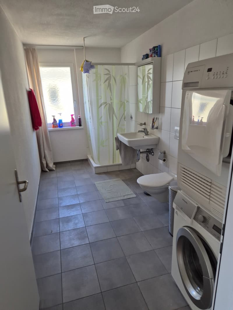 Modern bathroom with tiled floors, a sink, toilet, shower with bamboo-patterned curtain, mirror, washing machine, and cleaning supplies on the windowsill.