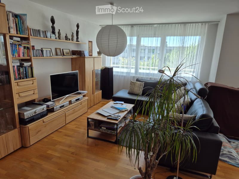 Modern living room with wooden flooring, large windows, a black leather sofa, a wooden coffee table, a bookshelf, and a television on a wooden stand.