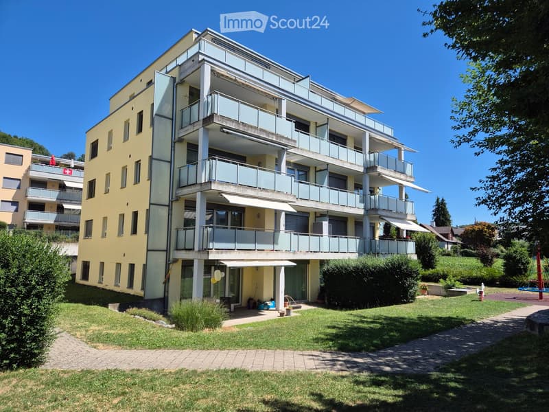 Modern multi-story residential building with balconies, surrounded by a well-maintained lawn and garden area.