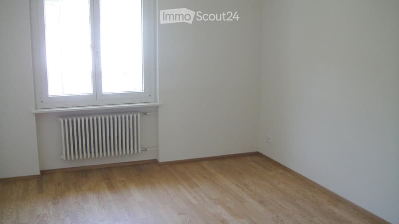 Empty room with wooden flooring, white walls, and a window with a radiator below it.