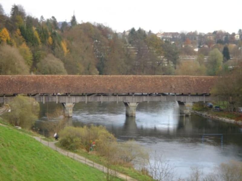 Scenic view of a wooden covered bridge spanning a river, surrounded by autumn-colored trees and a grassy bank.