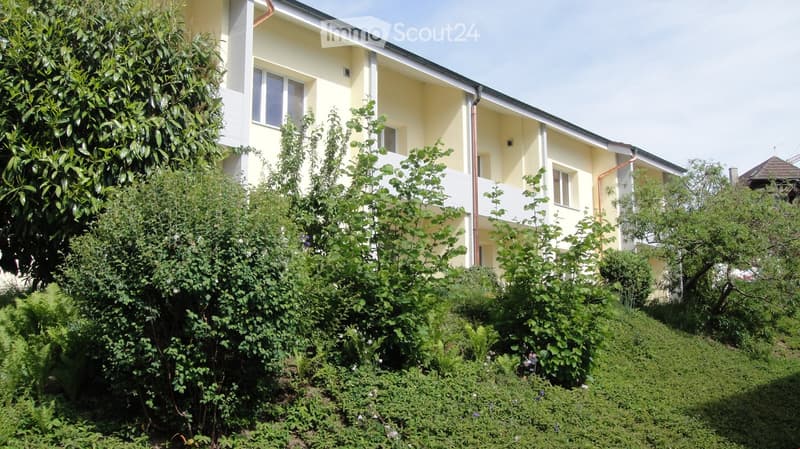 Two-story yellow building with white trim, multiple windows, and a small balcony on the second floor, surrounded by lush greenery and shrubs.