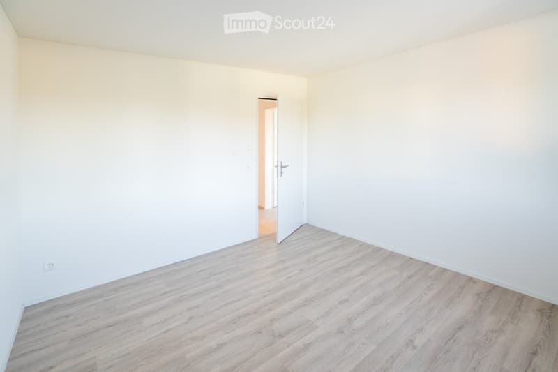 Empty room with light wooden flooring and white walls, featuring a partially open door leading to another space.