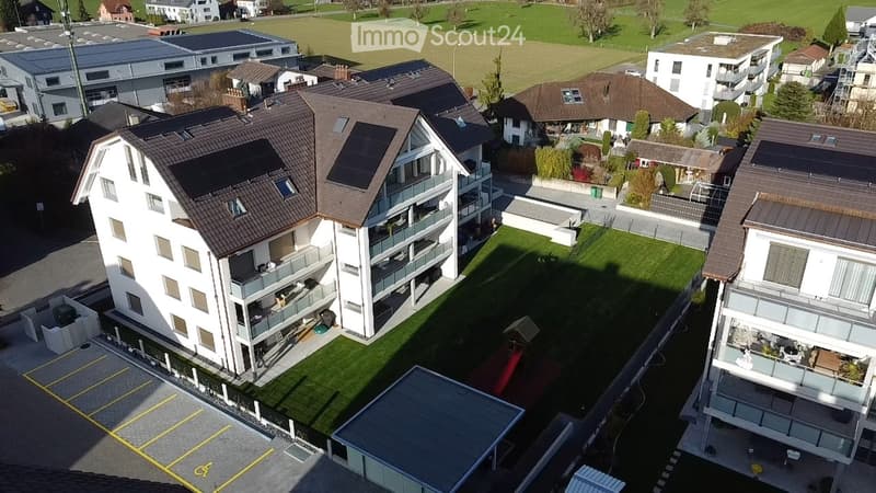 Aerial view of a multi-story residential building with balconies, surrounded by green lawns and other houses. Parking area visible in the foreground.